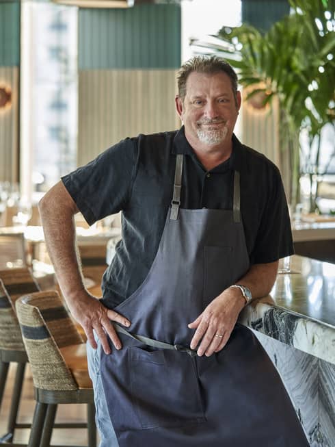 A chef in a black shirt and blue apron standing in a well lit restaurant.