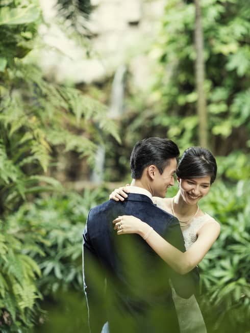 Smiling bride and groom embrace in garden with ferns, plants