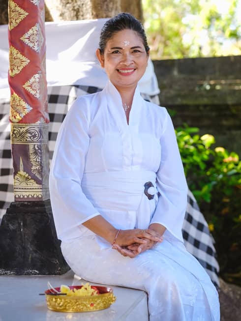 Smiling person in white linen outfit sits outdoors with greenery in background