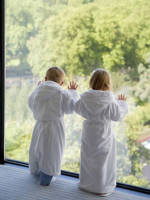Two young children wearing white bathrobes gaze out floor-to-ceiling window overlooking trees