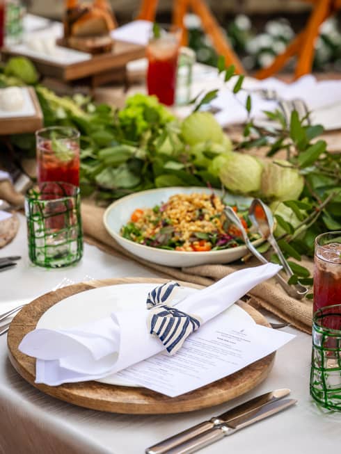 Place setting with a wooden charger, white plate, white napkin tied with a black-and-white striped ribbon sits in front of a large bowl of salad