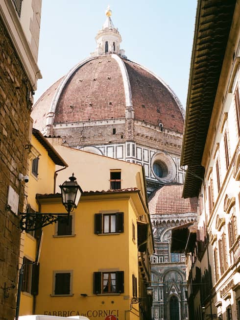 Looking up at the Florence Cathedral's dome from a narrow street below, a bright yellow shop building in the foreground.