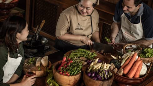 A chef teaching two participants in a cooking class, surrounded by fresh vegetables and ingredients in a rustic kitchen setting.
