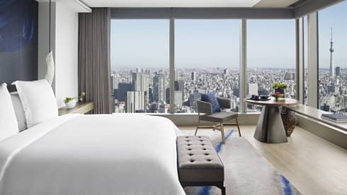 Guest room with king bed, tufted bench, round table with armchair and floor-to-ceiling windows offering city views