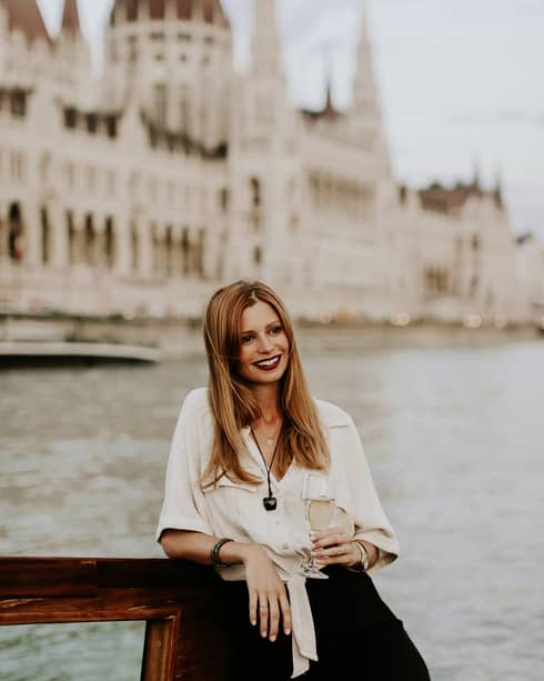 Long-haired women wearing ivory blouse and with glass of champagne in hand leaning on railing overlooking the Danube and Parliament building