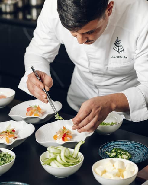 A chef adds garnishes to bowls of colourful food items in a food-prep area.