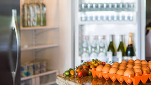Smiling man holds green bottle in front of open fridge, fresh eggs on counter