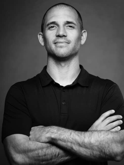 Black-and-white photo of man with buzzed hair cut wearing a black polo shirt and standing with his arms crossed