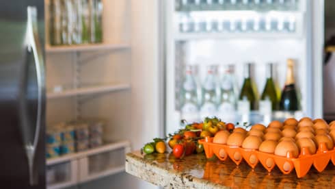 Smiling man holds green bottle in front of open fridge, fresh eggs on counter