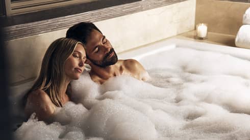 A couple sits together in a bubble bath with lit candles surrounding them, their heads leaning against eachother, both with their eyes closed