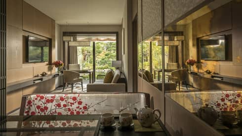 Connecting hotel room with sofa, desk and chairs, floral-patterned screen, tea set and large window overlooking a tranquil pond garden