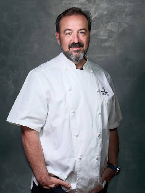 Smiling person in white double-breasted chef?s coat stands in front of gray background with hands in pockets