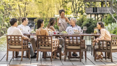 Group sharing a meal and drinks on an outdoor terrace with a chef grilling nearby, surrounded by lush greenery.