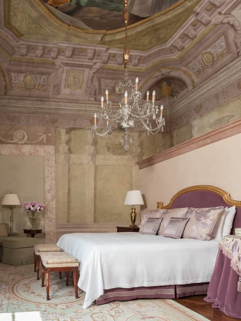 Frescoed Executive Suite bed with purple velvet headboard, nightstand with cloth under chandelier, vaulted ceiling
