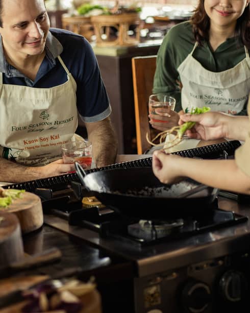 Three guests in aprons sit on stool in front of large wok, chef during Thai cooking class