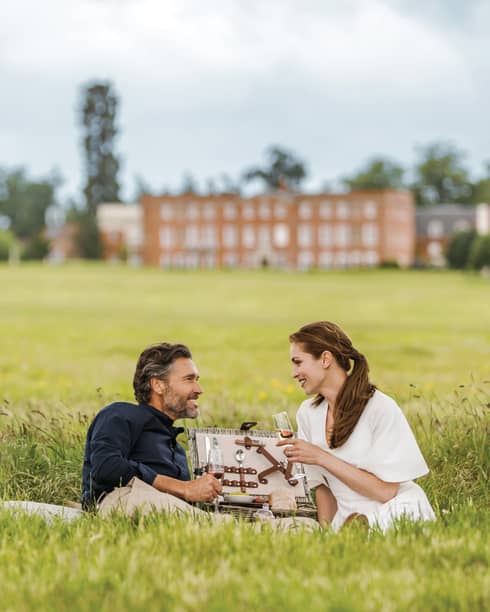 Couple with wine glasses lies in tall green grass with open picnic basket, hotel manor in background