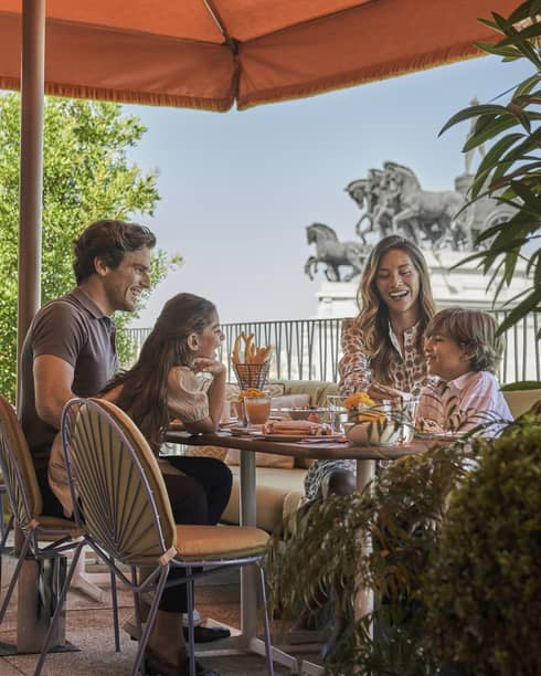 Family enjoying breakfast outdoors under an orange canopy, with views of horse statues in the background