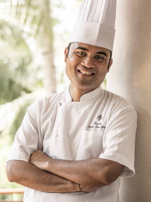 Smiling person in white chef's jacket and hat stands with arms crossed on balcony