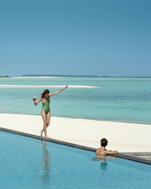 A guest wades in a pool on a beach overlooking the ocean as another guest balances on the edge with a drink in hand.