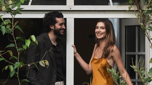 Two people stand on a vine-covered balcony surrounded by foliage.