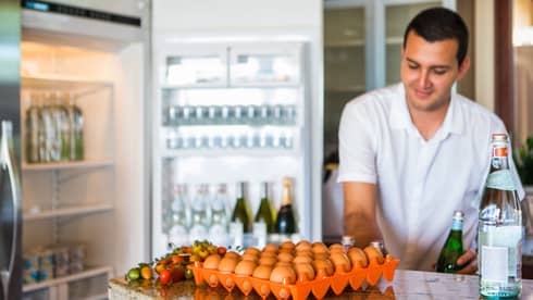 Smiling man holds green bottle in front of open fridge, fresh eggs on counter