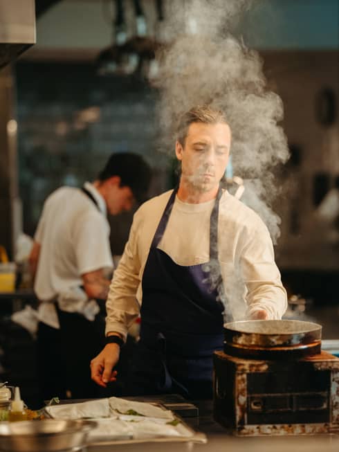 Chef wearing white long-sleeve shirt and dark-blue apron tends to a pot with rising steam