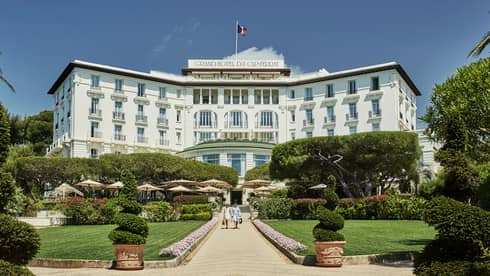Two people walk down sunny stone path under large Grand-Hotel du Cap-Ferrat