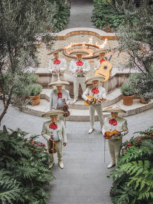 A group of mariachi musicians in an outdoor courtyard with paved pathways, abundant greenery and a fountain with a flame feature in the middle of it