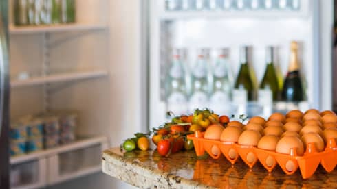 Smiling man holds green bottle in front of open fridge, fresh eggs on counter