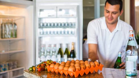 Smiling man holds green bottle in front of open fridge, fresh eggs on counter