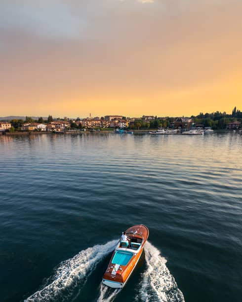 Person driving a wooden speedboat across a lake towards shore at sunset