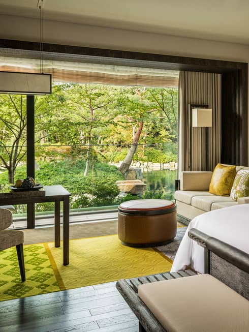 Kyoto accommodation suite room with king bed, green-cushioned sofa, round ottoman, and large window overlooking a pond garden.