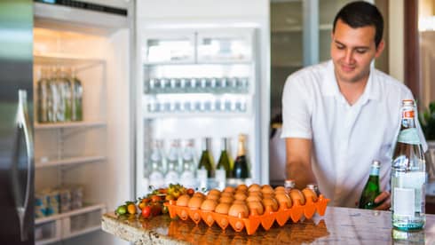 Smiling man holds green bottle in front of open fridge, fresh eggs on counter