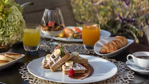 A table filled with breakfast items including sliced waffles topped with fruit, a croissant, two cups of juice, cup of coffee and a glass of yogurt parfait