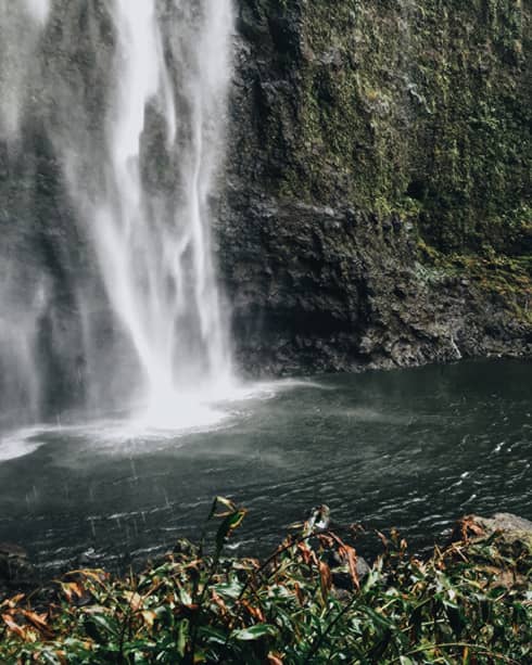 Back of woman hiking over black rocks under tall waterfall, small pond