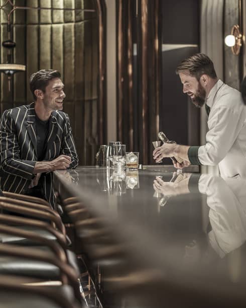 Bartender preparing a drink while a customer sits at the bar, both smiling, in a modern, upscale bar with a reflective countertop and warm lighting.