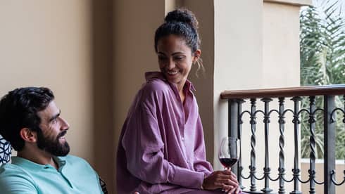 Man and woman, wine glasses in hand, smile at each other while sitting on balcony facing blue sky