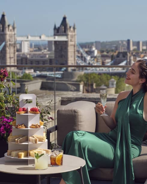 Woman in green dress holding champagne beside tea stand with confections and Tower Bridge in backdrop