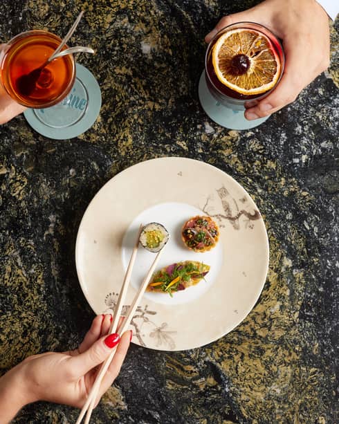 Left hand holding cocktail and right hand picking up sushi with chopsticks