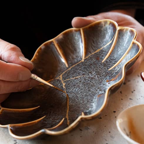 A person using the art of Kintsugi, by using gold paint to paint over the cracks of a once broken black flower shaped plate.