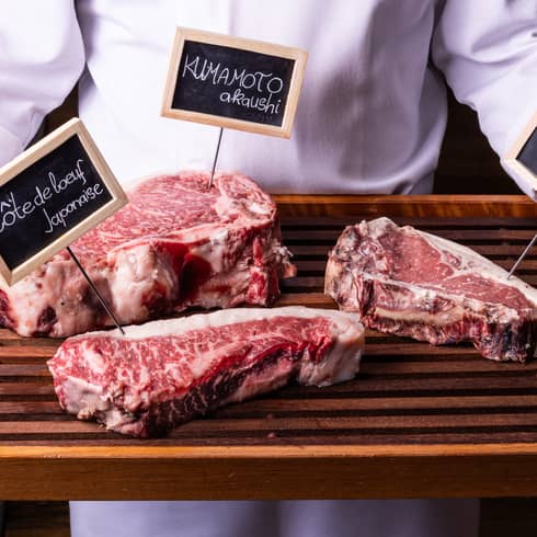 Close-up view of a wood tray with three labelled cuts of well-marbled beef, held by person in white chef's coat.