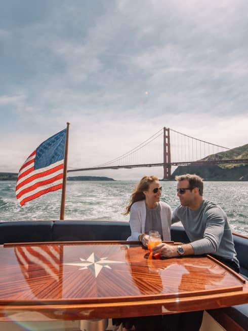 A man and woman on the back of a boat with a metal bridge in the distance behind them and an American flag on the boat.