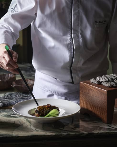 Chef using chopsticks to plate black pork belly in a white dish near a dozen black spiky delicacies on a small wood platform.