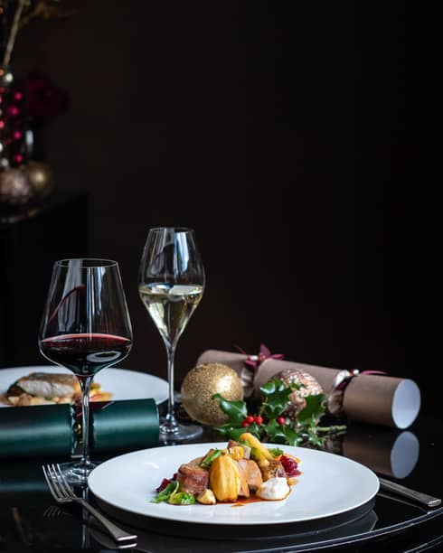 A festive meal on round white plate with two glasses of wine, one red and one white, and holly berry garnishes
