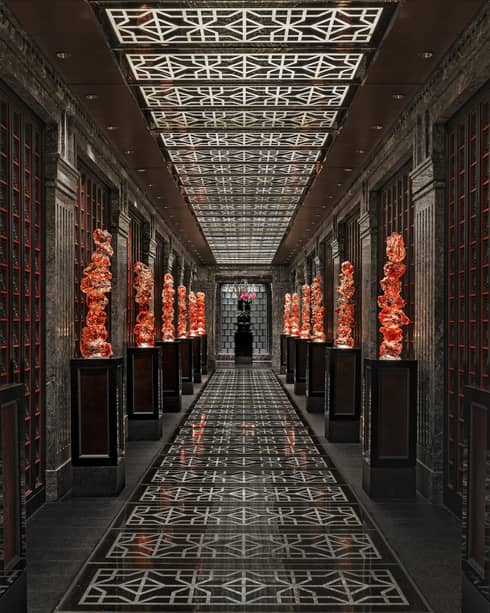 View down a long black-tiled hall lined with orange glass sculptures on pedestals, the geometric ceiling mirroring the floor.