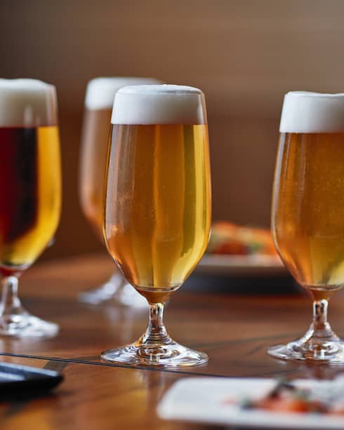 Four tall beer glasses filled with beer on a table with cheese, crackers and appetizers