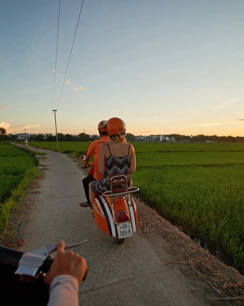 Two people on a Vespa head toward the distant sunset, along a narrow path bordered by green fields and distant houses.