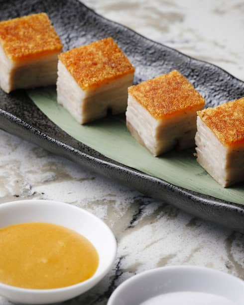 Cubes of crispy pork belly on elongated plate with dipping sauce in nearby bowl