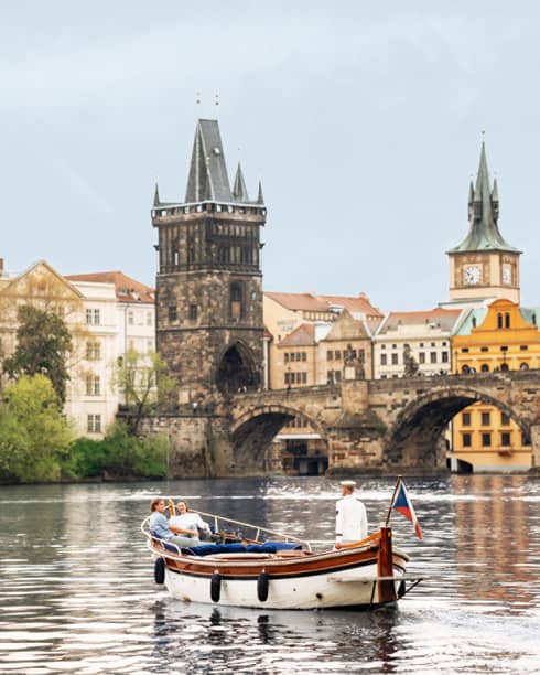 Couple in wooden boat with captain on Vltava River with baroque buildings in backdrop