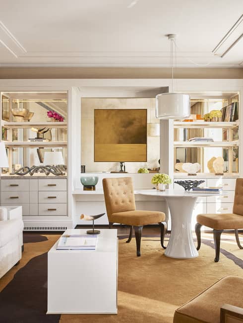 Luxury hotel living room with plush seating, tan chairs, round and rectangular tables, TV, decorative shelves and large windows with beige curtains